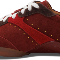 A red suede and leather sneaker with white laces, brown heel tab, and gold-tone decorative studs, shown from a side profile on a white background.