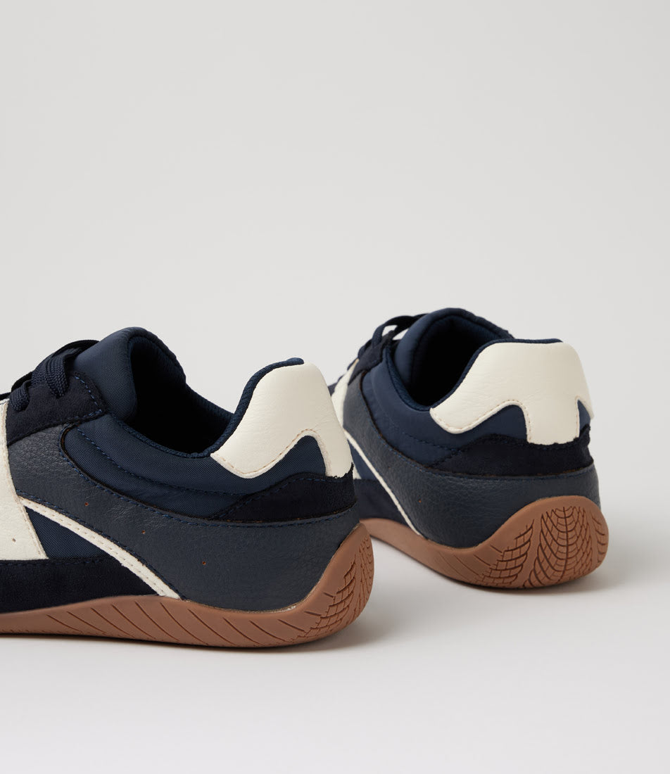 A pair of low-top sneakers shown from the rear three-quarter angle, featuring navy blue and off-white leather uppers with a brown gum sole, set against a plain light gray background.