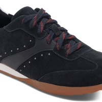 A low-top sneaker in black suede and synthetic leather, featuring black laces with red stripes, a white midsole, and a brown gum outsole, shown on a white background.